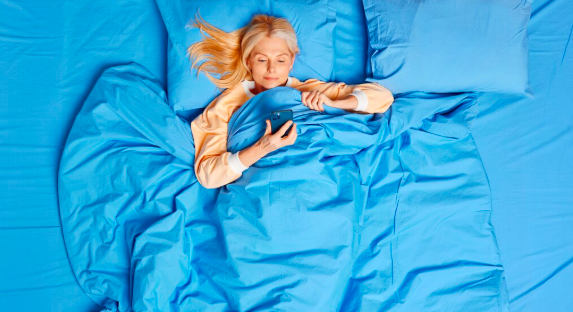 Powyżej widok poważnej blondynki w średnim wieku ma uzależnienie od gadżetów trzyma smartfon leży w wygodnym łóżku sprawdza konto w serwisie społecznościowym przed zaśnięciem czyta wiadomości online będąc sam